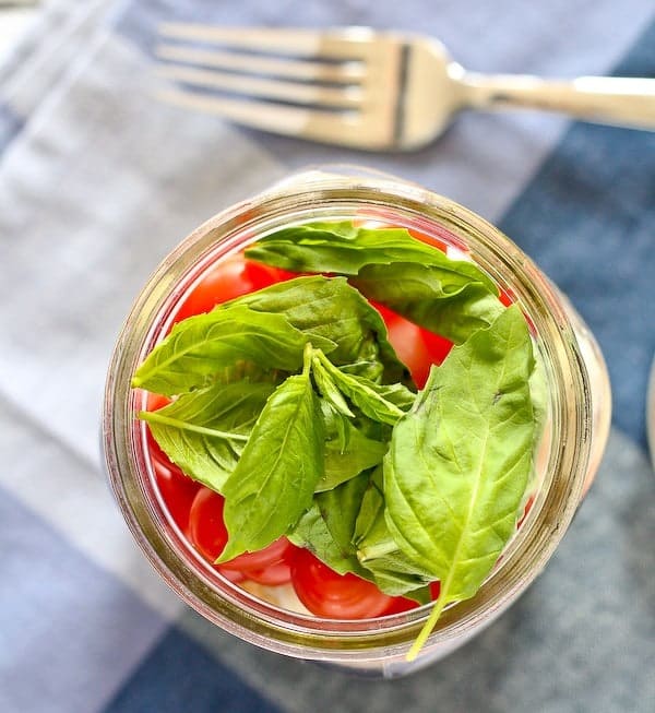 Top view of Mason jar picturing several basil leaves on top of grape tomatoes. Fork and blue plaid linens are in background.