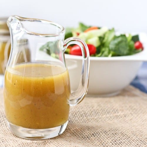 Front view of small clear glass pitcher containing honey mustard vinaigrette, with salad in background.
