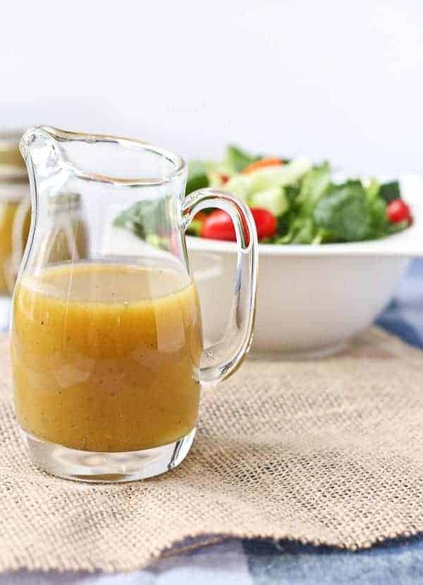 Front view of small clear glass pitcher containing honey mustard vinaigrette, with salad in background.