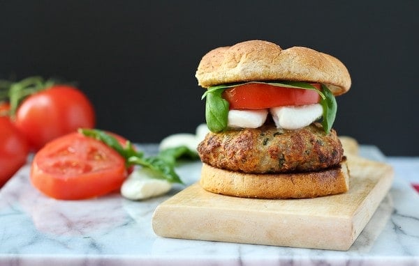 Front view of burger on board, arranged with sliced tomato, fresh mozzarella, and basil on marble cutting board.