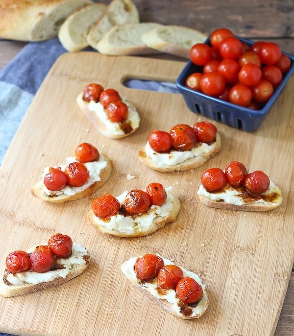 Overhead of several crostini arranged on cutting board, with container of fresh cherry tomatoes and sliced bread.