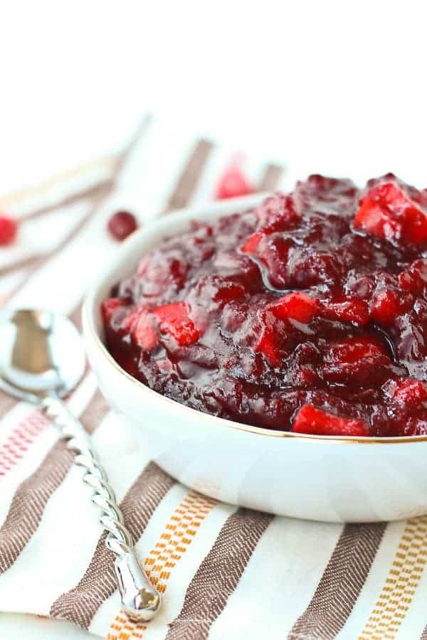 Partial front view of cranberry sauce in white bowl with gold rim, with spoon along side and scattered cranberries on striped cloth.