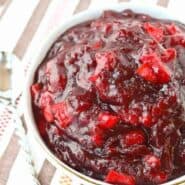 Overhead of white bowl filled to the brim with cranberry sauce, on tan and white striped cloth.