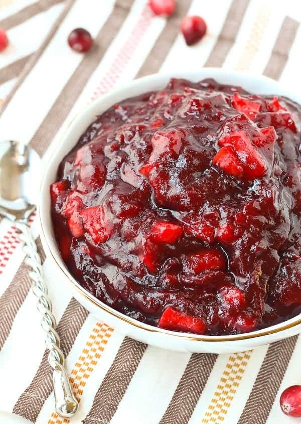 Overhead of white bowl filled to the brim with cranberry sauce, on tan and white striped cloth.