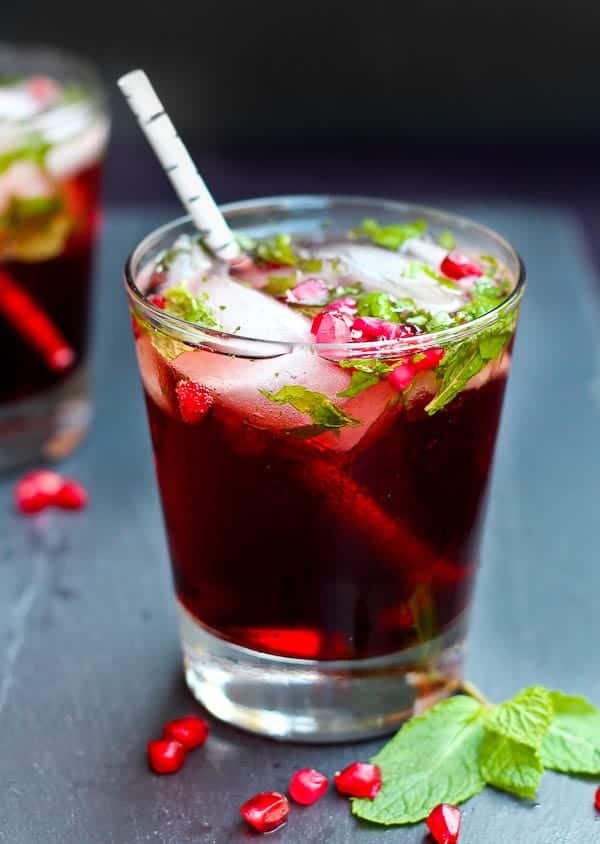 Front view of clear glass with pomegranate mojito, with white "birch" straw. On black background with mint leaves and pomegranate arils scattered around glass.