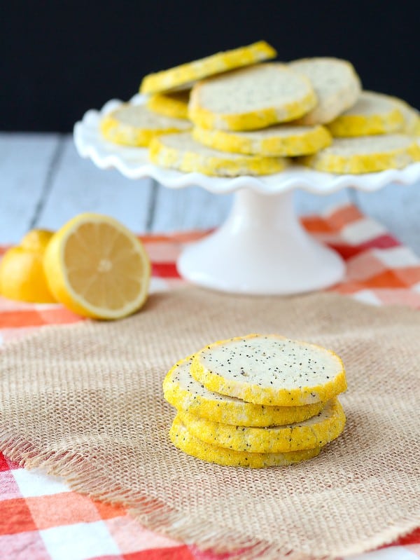 Four poppy seed cookies stacked on burlap, with cut lemon and pedestal dish holding more cookies in background.