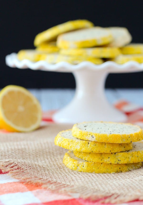 A stack of four poppy seed cookies in the foreground, and more cookies on a white pedestal dish in the background.