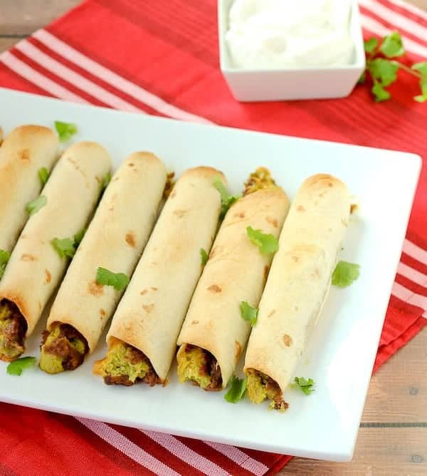 Overhead of baked taquitos lined up on white rectangular platter, garnished with cilantro, on red striped cloth.