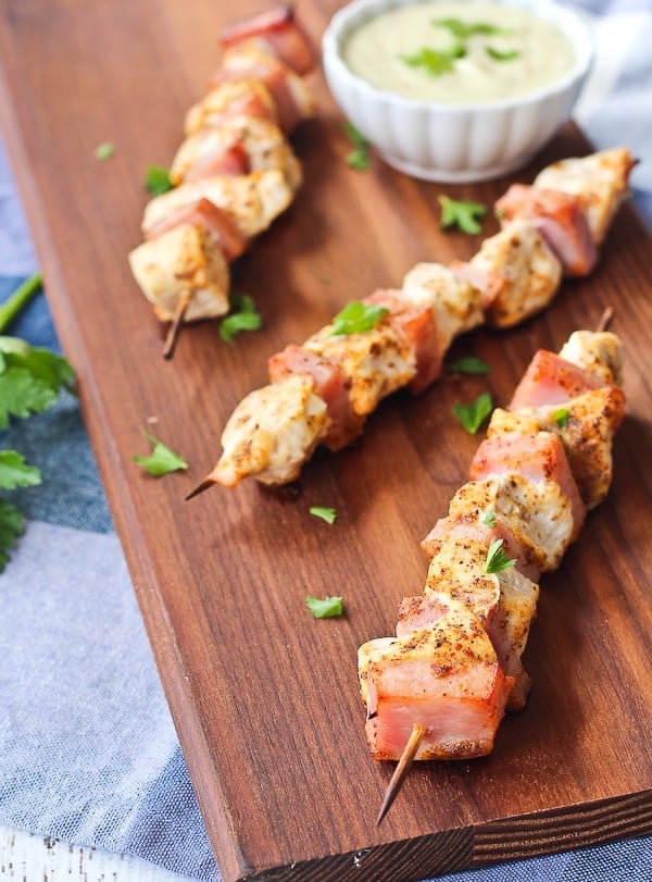 Overhead of three kabobs on wooden cutting board with sauce in small white bowl.