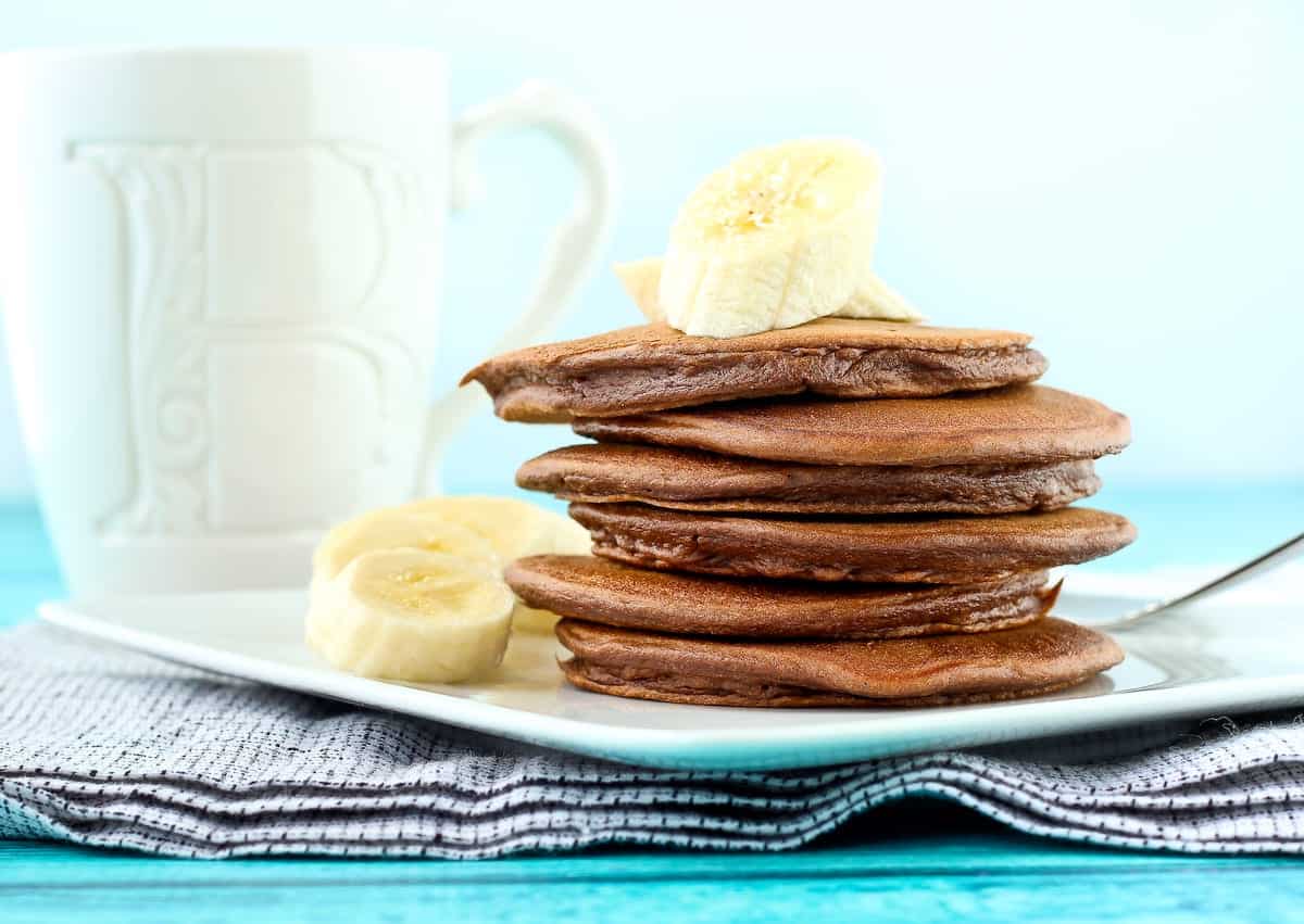 Stack of six chocolate pancakes on plate with sliced banana, pitcher in background.