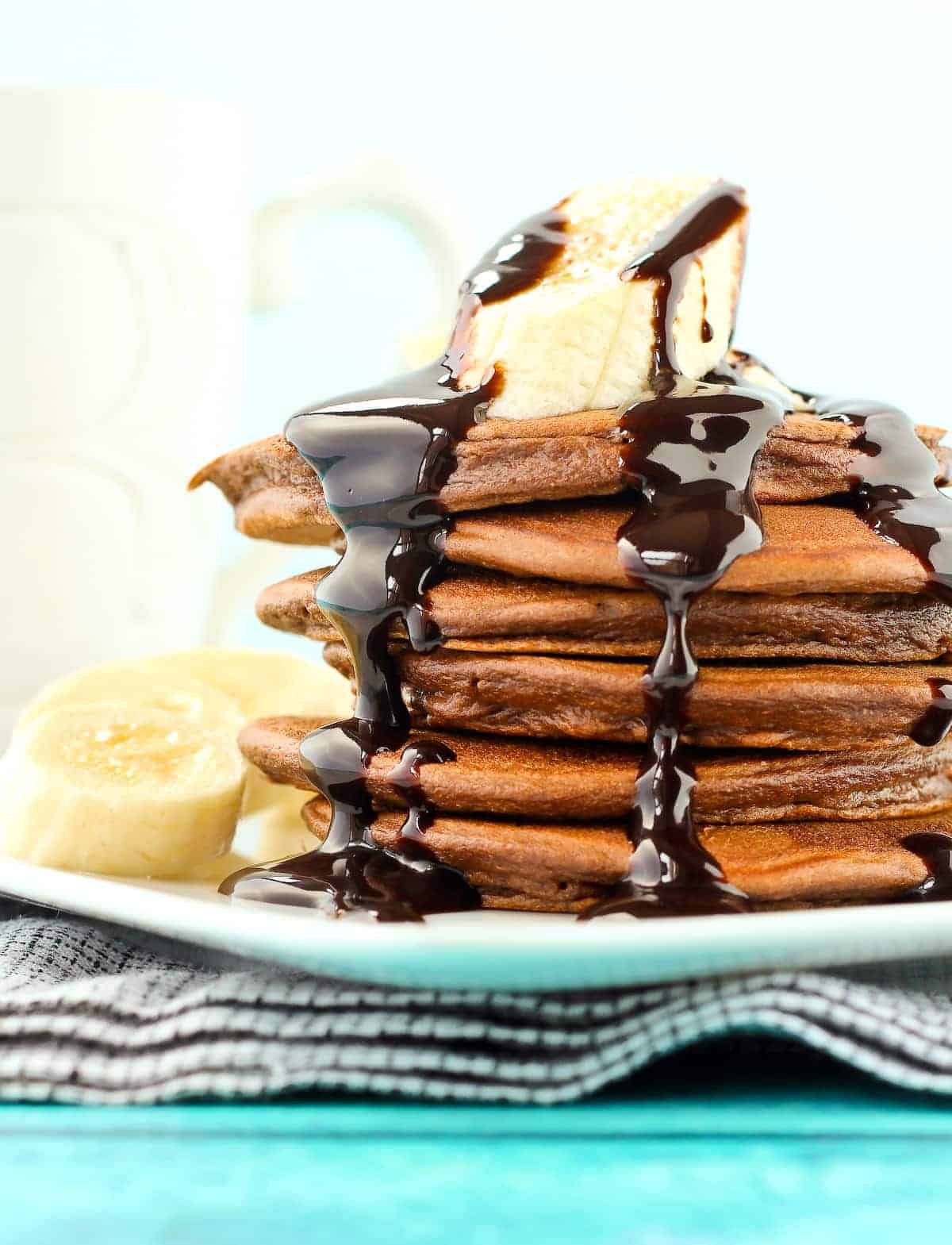 Six chocolate pancakes stacked on plate, with sliced banana and chocolate syrup.