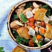 Overhead of earthenware bowl containing sausage, bean, and kale soup garnished with large croutons.