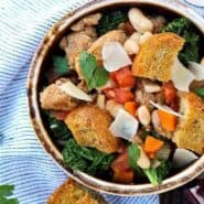 Overhead of earthenware bowl containing sausage, bean, and kale soup garnished with large croutons.