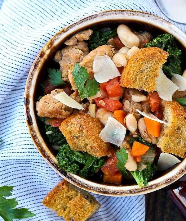 Overhead of earthenware bowl containing Italian sausage stew garnished with croutons, Parmesan, and fresh parsley.