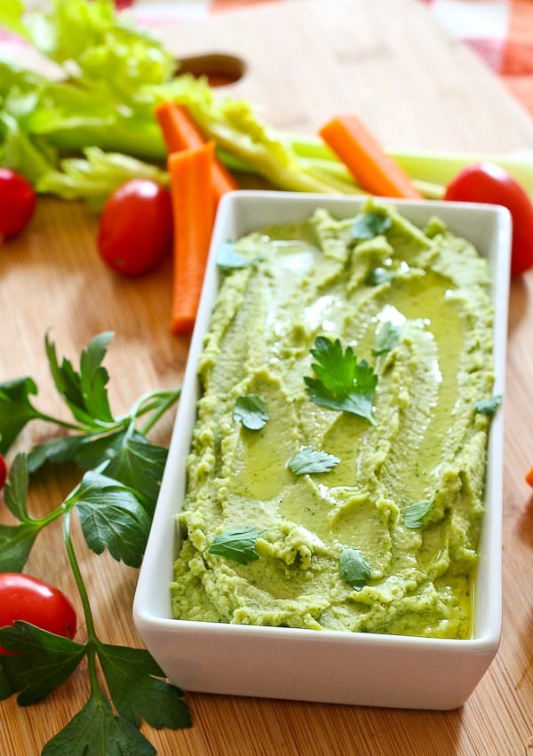 White rectangular serving dish with hummus, garnished with parsley leaves, surrounded by fresh veggies.