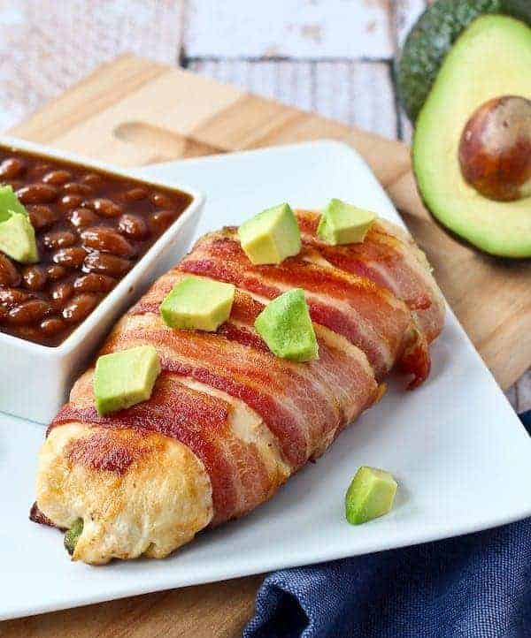 Chicken breast on square white plate with bowl of baked beans, garnished with cubes of avocado.