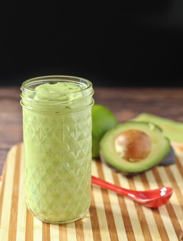 Small clear jar containing avocado dressing on striped bamboo board, with red spoon and cut avocado in background.