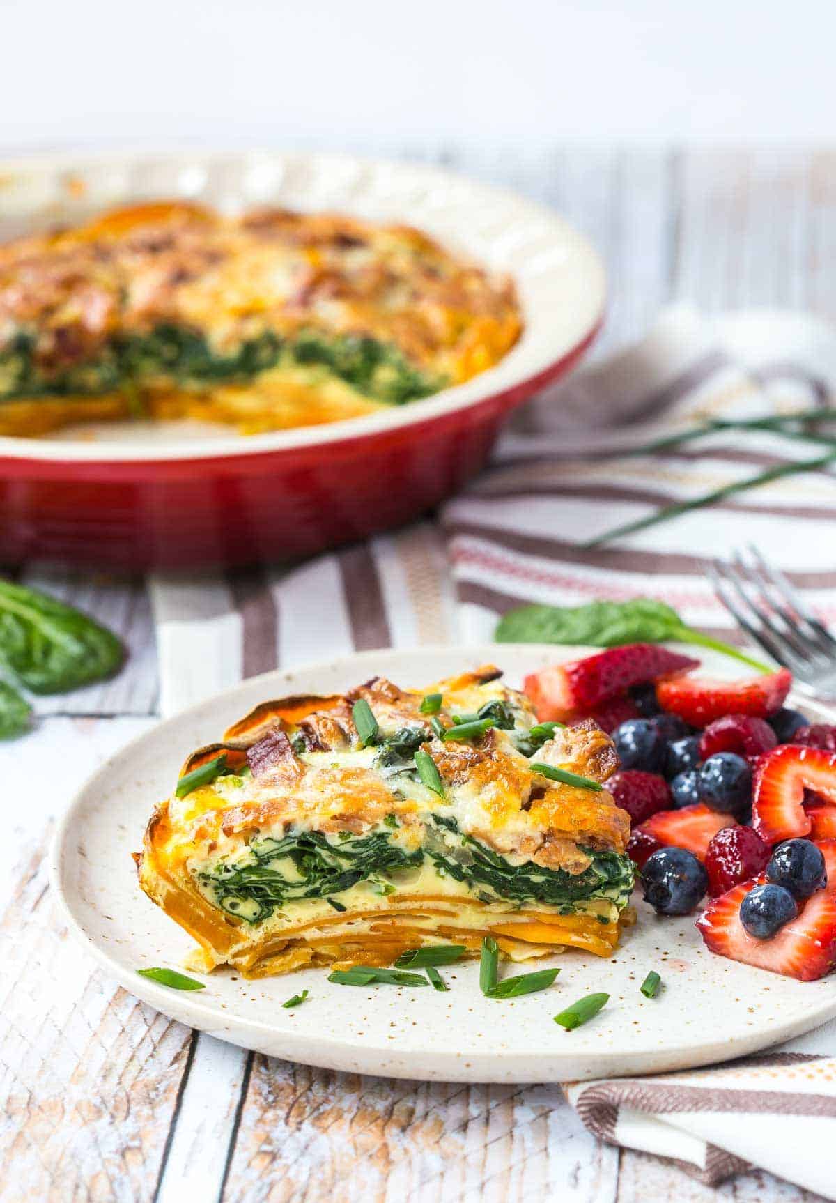 A slice of quiche on round white plate garnished with chopped chives, with a side of fresh berries.