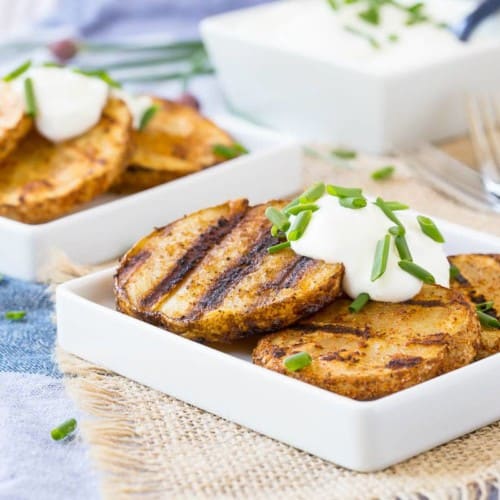 Grilled potatoes on square white plates, garnished with sour cream and chopped chives.