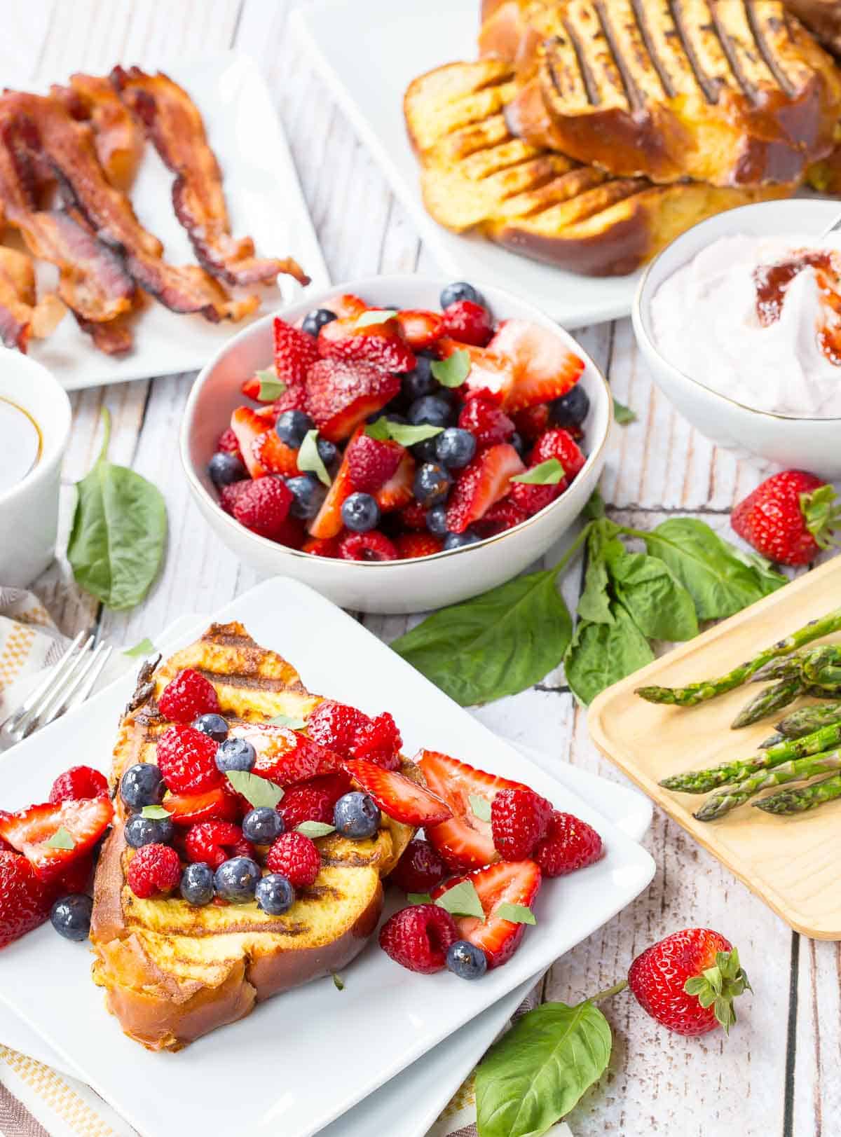 Slices of grilled French toast on a square plate garnished with mixed berries, next to a bowl of berries, a plate of bacon, and more French toast in the background.