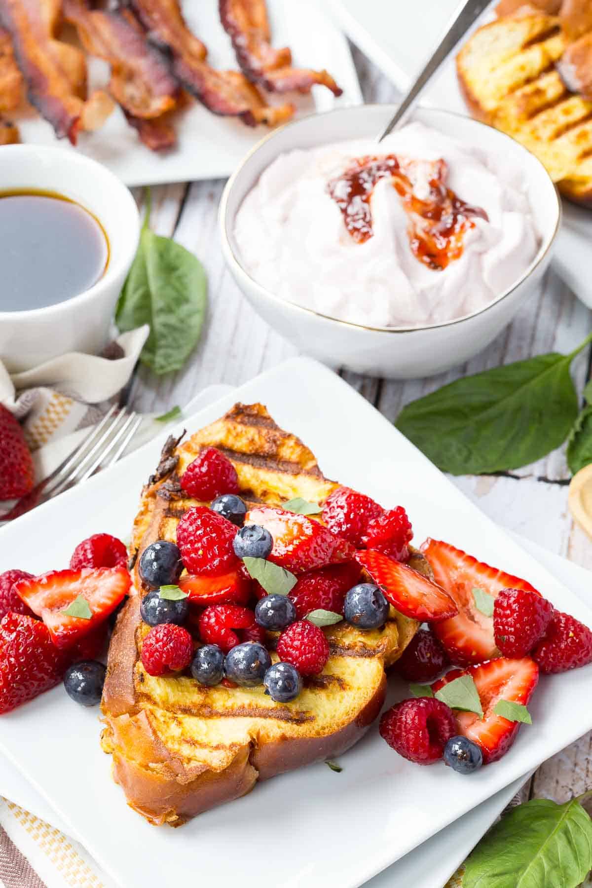 Slices of grilled French toast on a square plate garnished with mixed berries, next to a bowl of yogurt, a plate of bacon, and more French toast in the background.