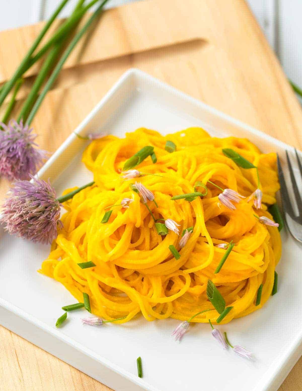 A serving of pasta mixed with carrot pesto, garnished with chives, in shallow square white dish.