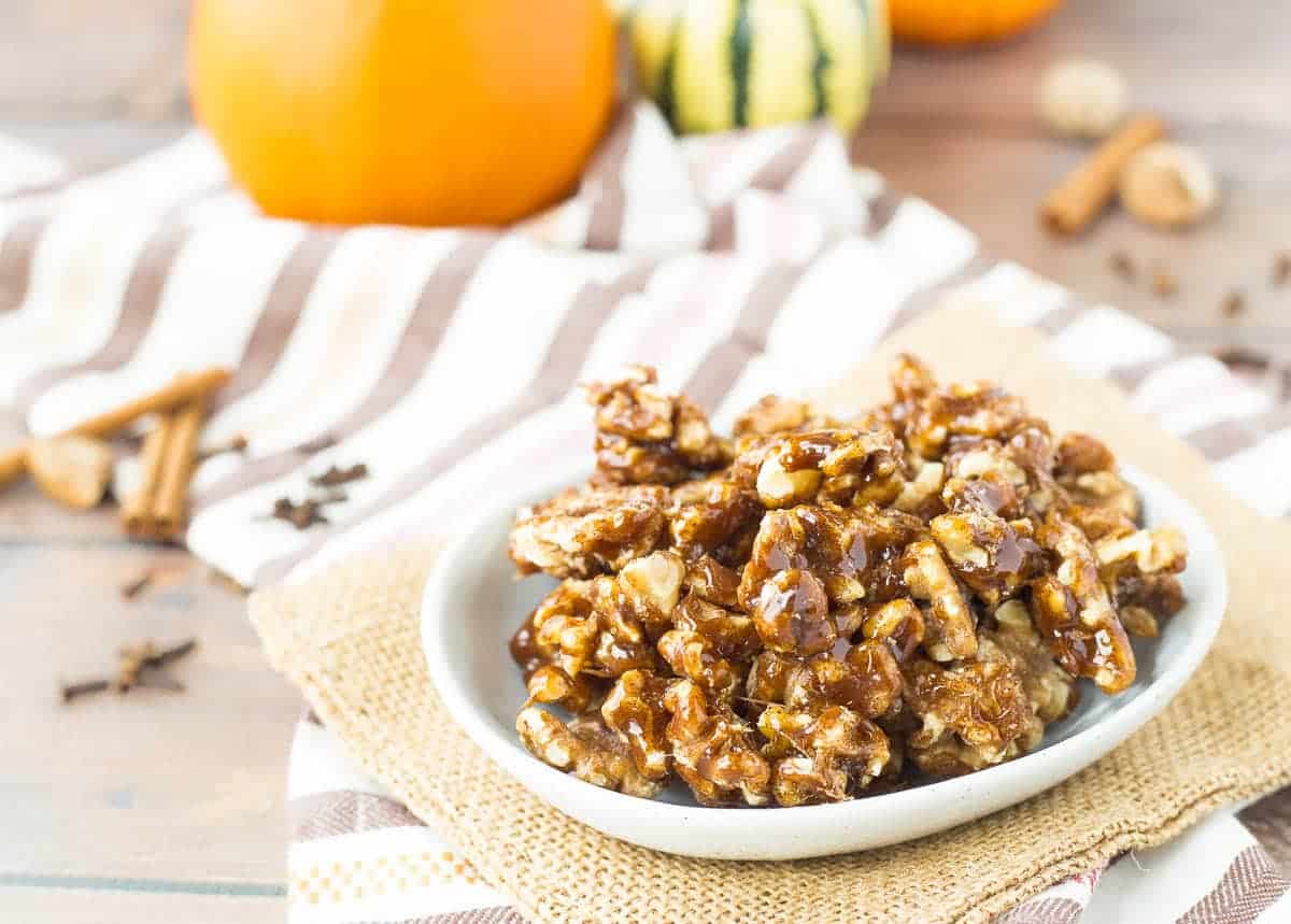 A bowl of pumpkin spice candied walnuts on a table with orange pumpkins in the background.