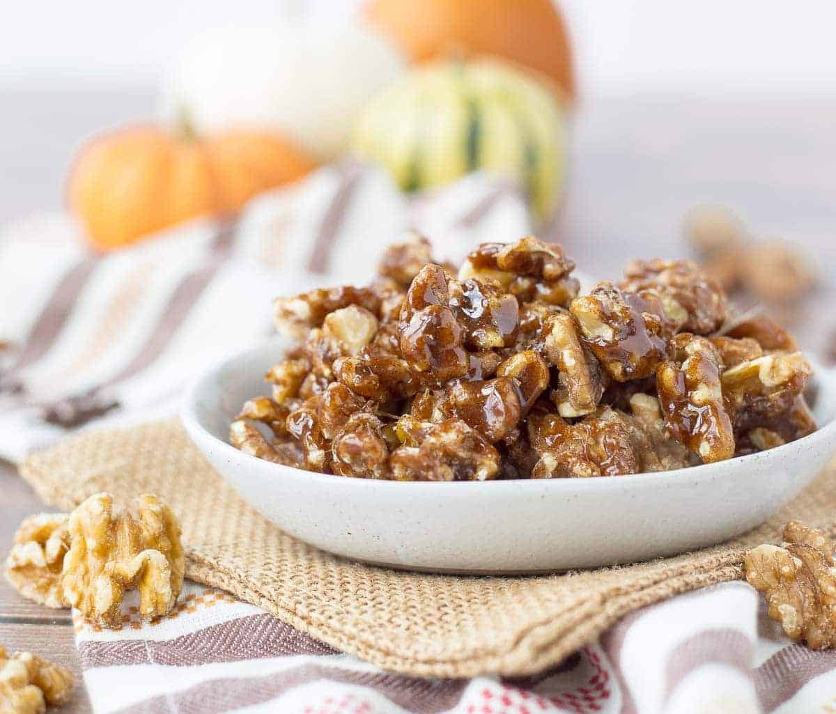 A bowl of pumpkin spice candied walnuts on a table with orange pumpkins in the background.