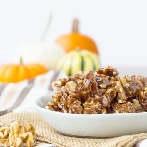 A bowl of pumpkin spice candied walnuts on a table with orange pumpkins in the background.