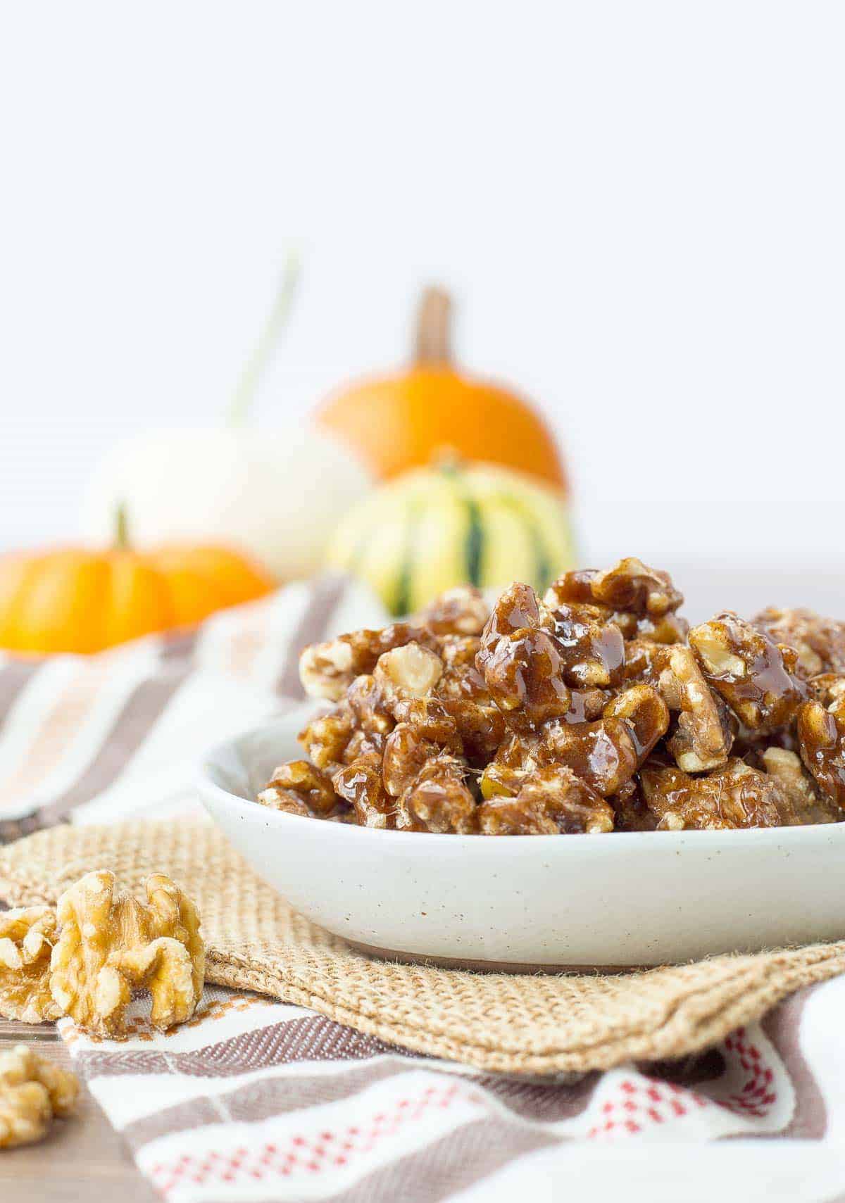 A bowl of pumpkin spice candied walnuts on a table with orange pumpkins in the background.