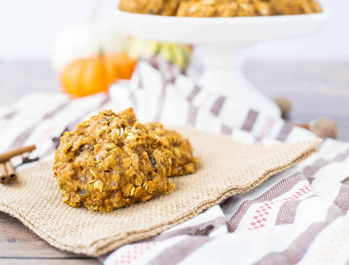 Two breakfast pumpkin cookies on a burlap napkin, with more cookies on a cake stand in the background.