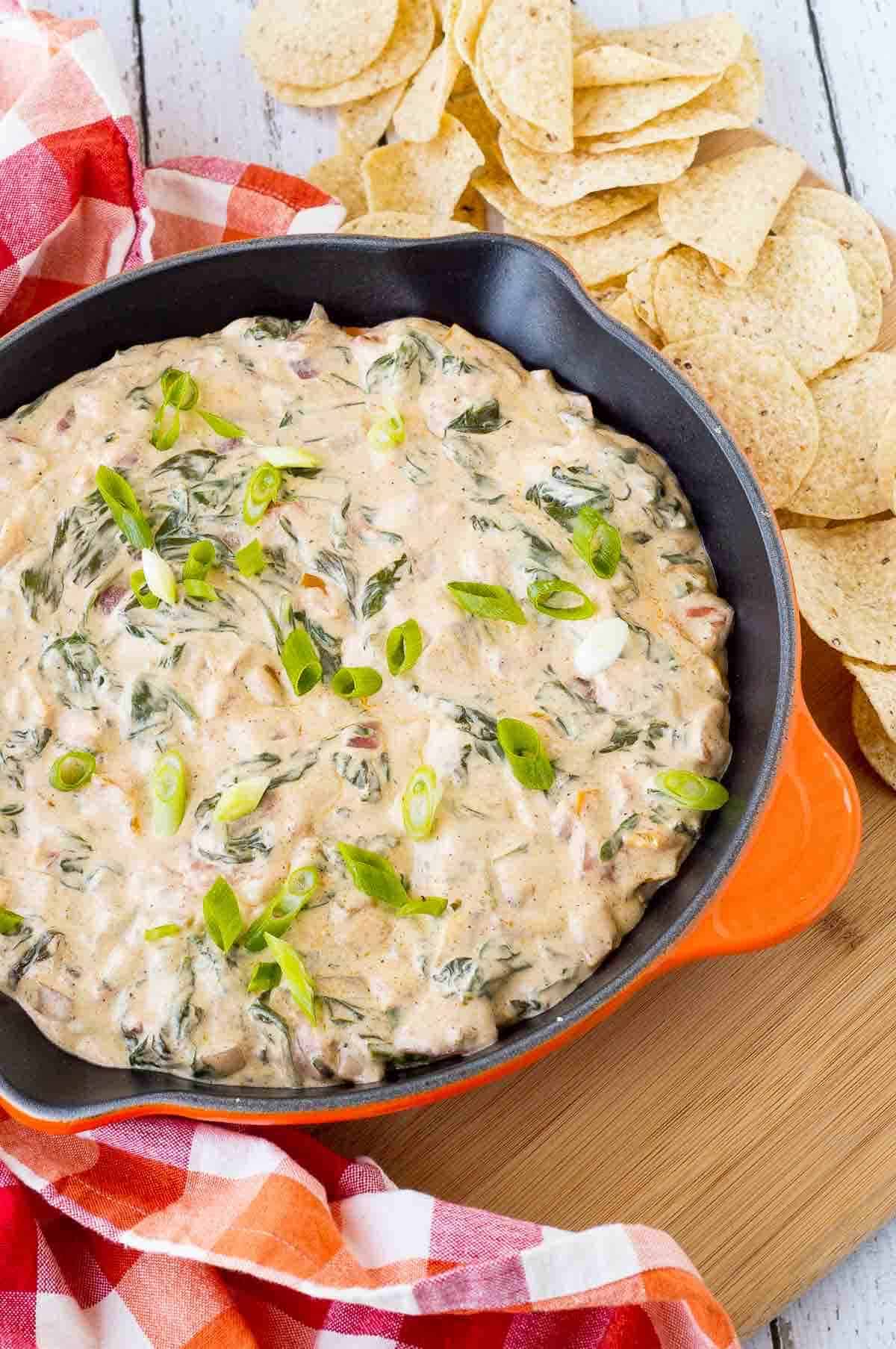 Overhead of spinach dip in small orange cast iron skillet, surrounded by tortilla chips and red plaid cloth.