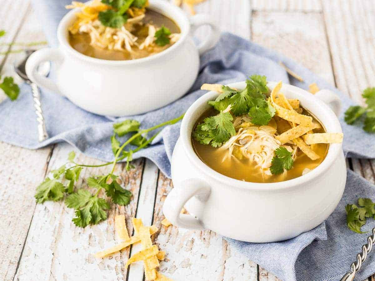 Chicken enchilada soup in bowls, on blue denim cloth, with toppings scattered around.