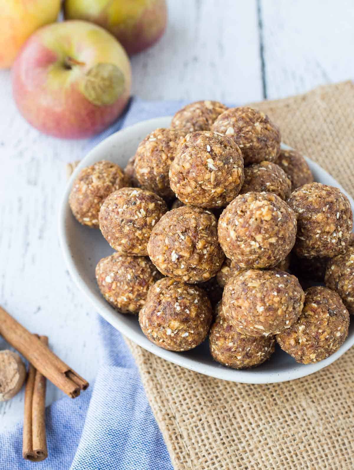 Several energy bites in oval white dish, with apples and cinnamon sticks in background.