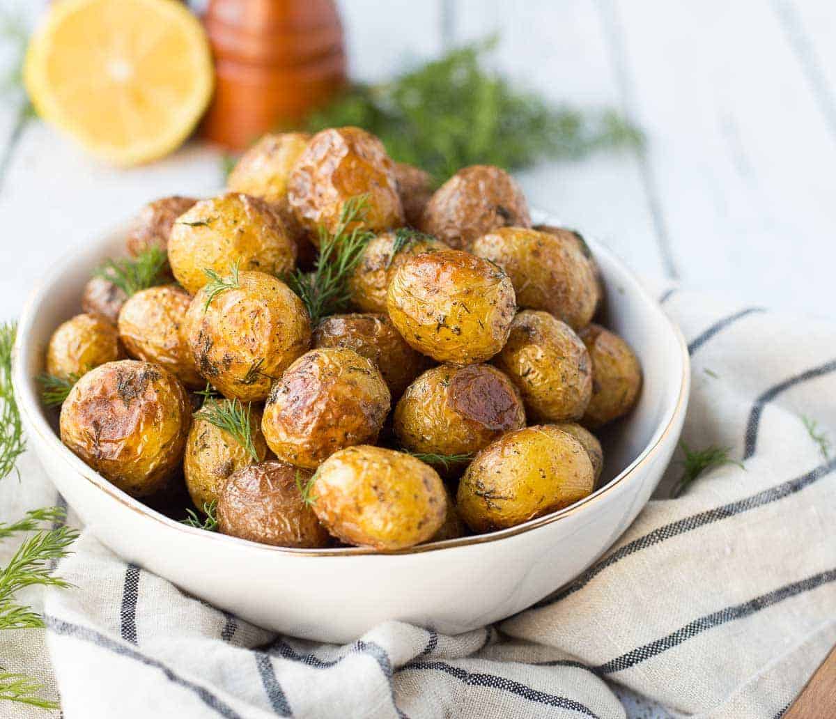 Front view of crispy roasted potatoes, on striped cloth, with dill, lemon, and pepper grinder in background.