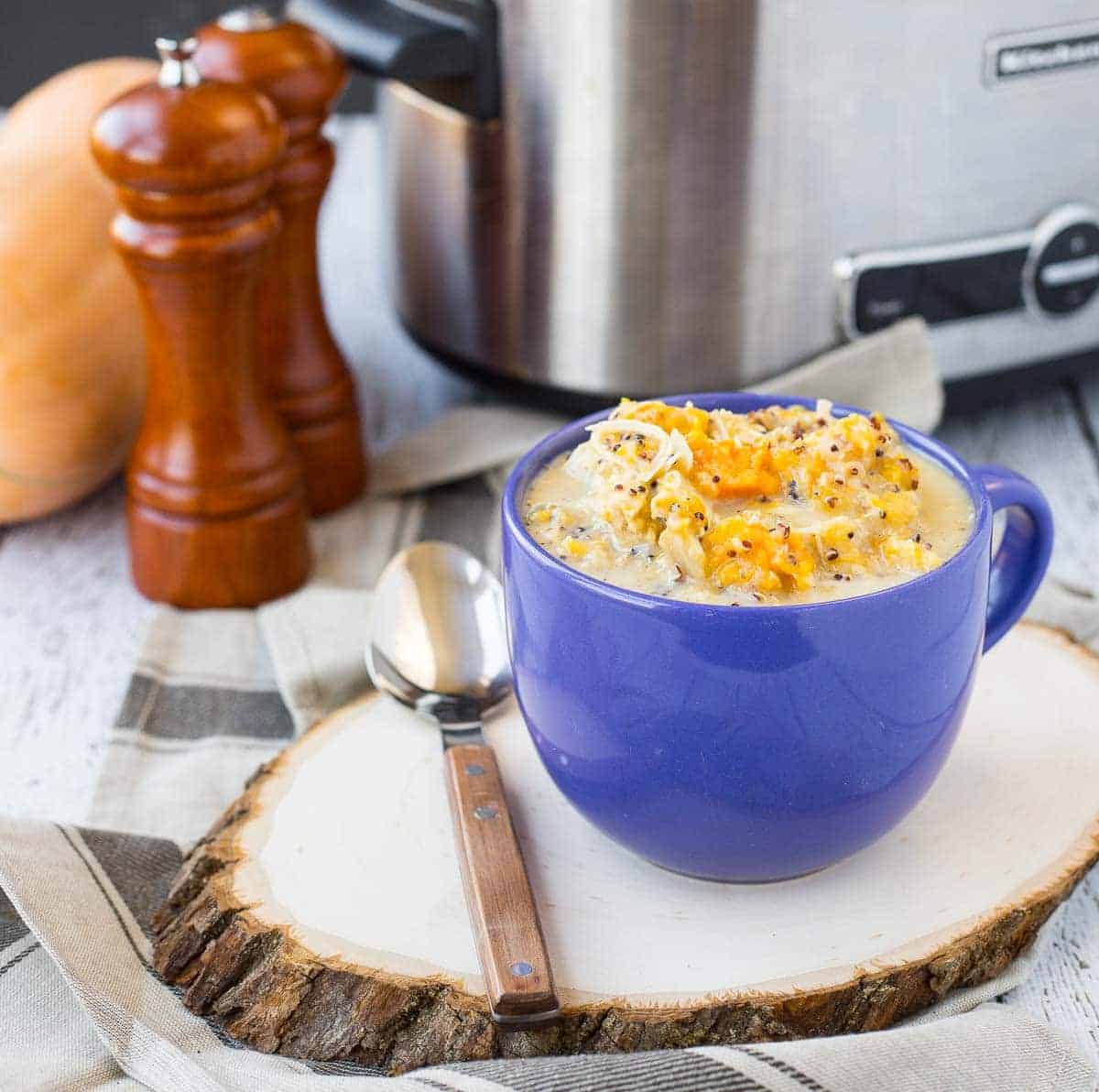 Soup in blue mug with crockpot, squash, and pepper/salt mills in background.