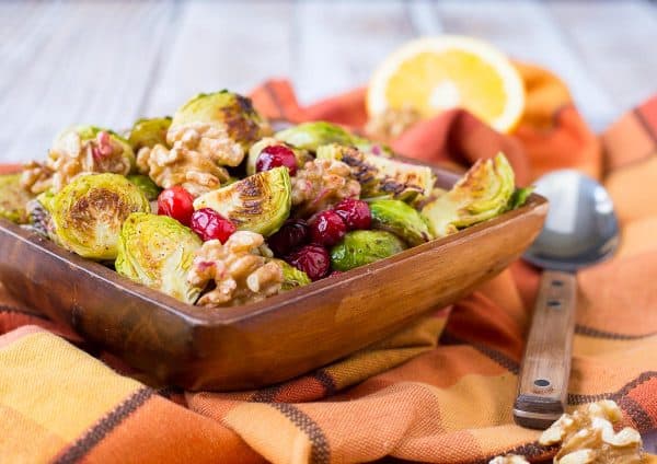 Front view of wooden bowl with brussels sprouts, along with spoon, walnuts halves and orange half.