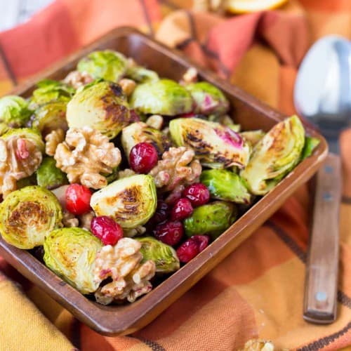Overhead of roasted brussels sprouts in square wooden dish, with spoon along side, on orange plaid cloth.