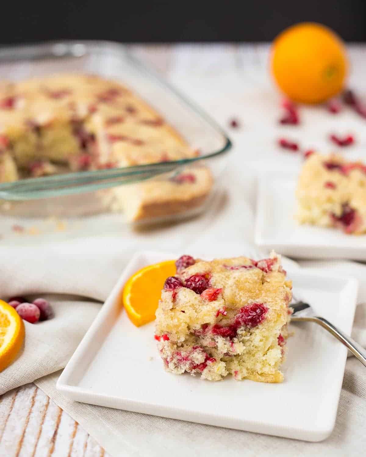 A piece of cranberry coffee cake on square white plate with fork, garnished with a half moon orange slice.