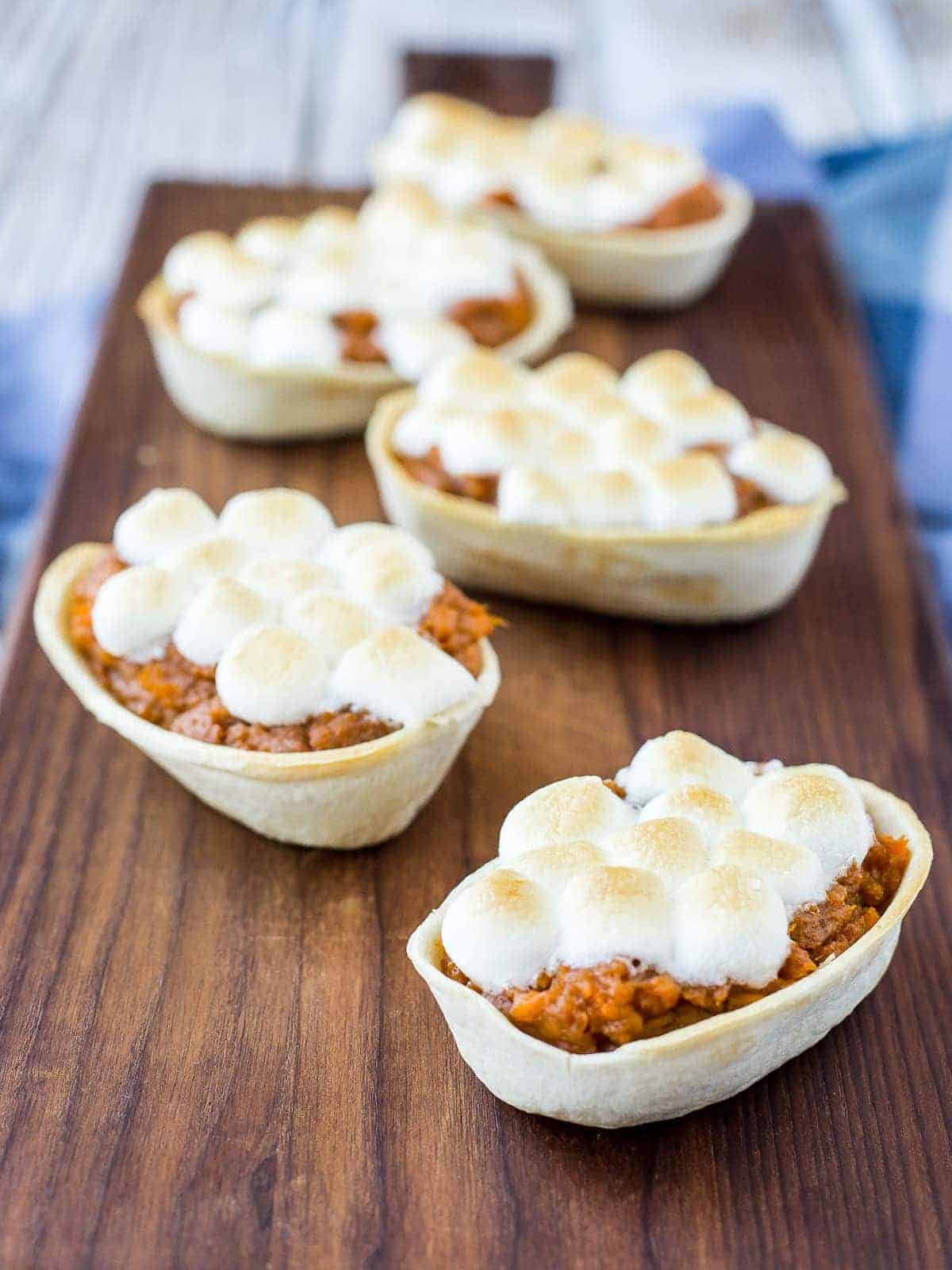 Five mini boats filled with sweet potato, topped with marshmallow, arranged on a long brown board.