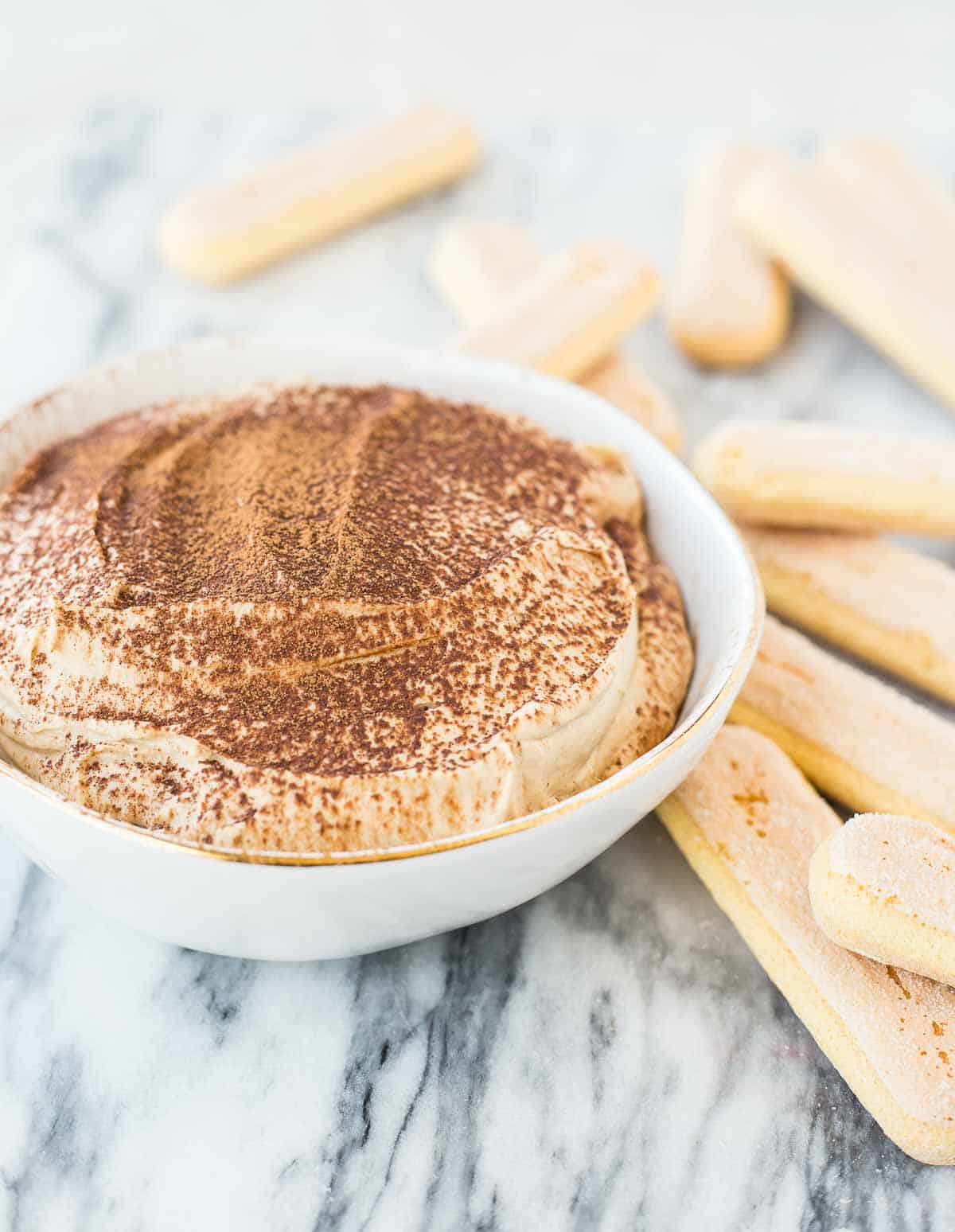 Closeup of tiramisu dip in a round white bowl, garnished with cocoa powder, and surrounded by ladyfingers, on a white marble surface.
