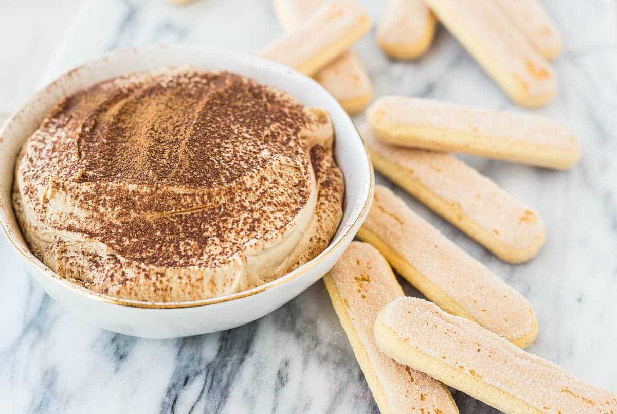 Tiramisu dip in small white bowl, garnished with a dusting of cocoa powder, with lady fingers arranged around it.