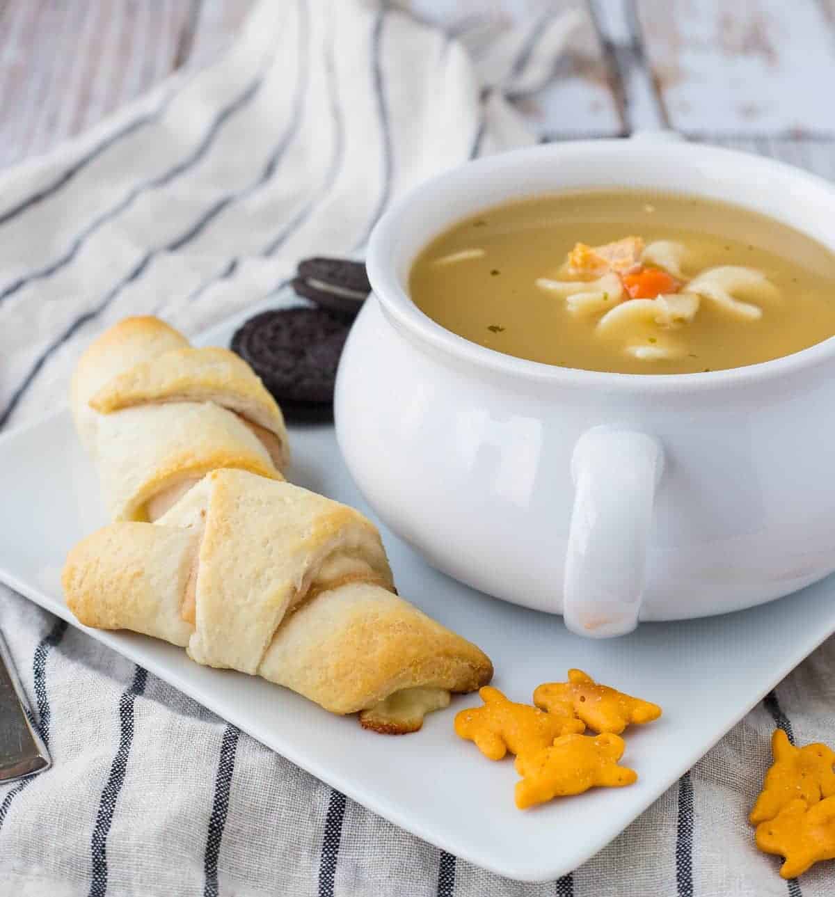 Soup in white bowl, on square white plate with turkey crescent roll sandwich.