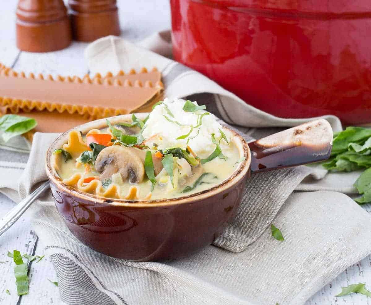 A serving of vegetarian lasagna soup in brown bowl with short handle. Red dutch oven and uncooked lasagna noodles in background.