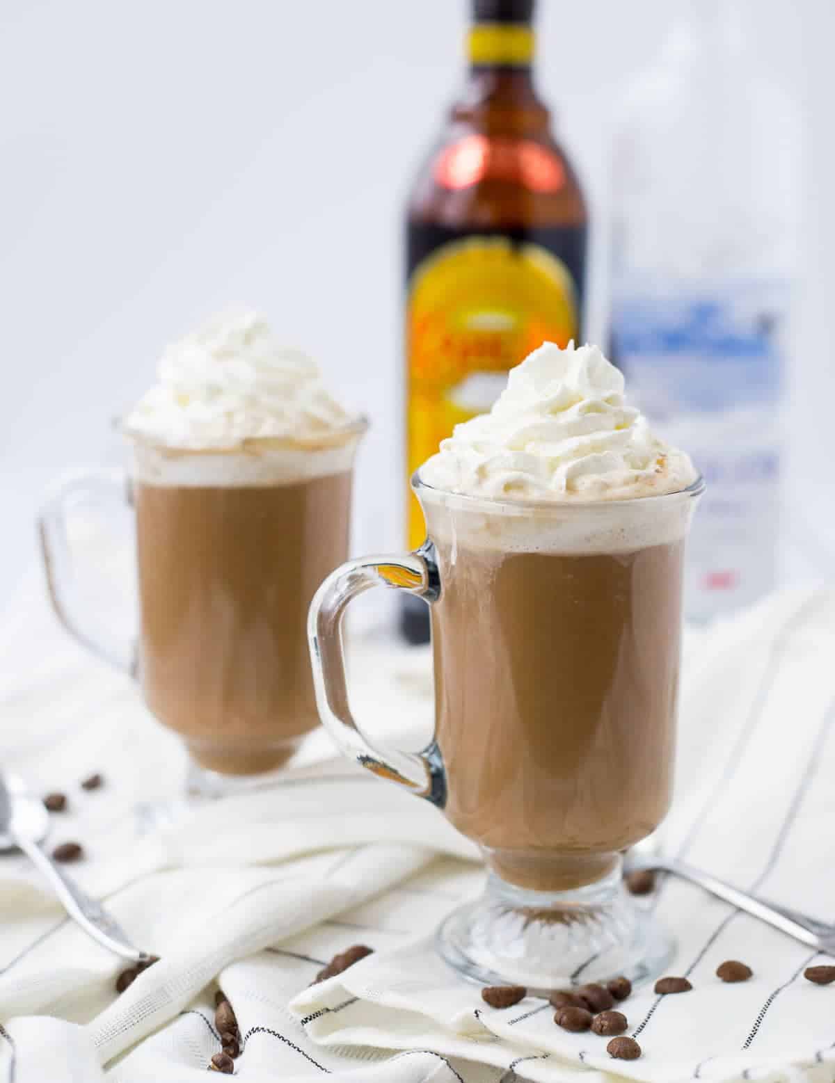 Footed clear glass mugs containing hot chocolate garnished with whipped cream, on white striped cloth, with bottle of Kahlua in background.