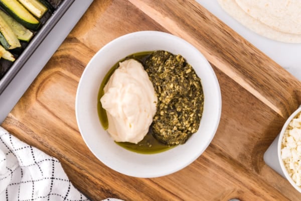 Mayo and pesto combined in a white bowl on a wooden cutting board.