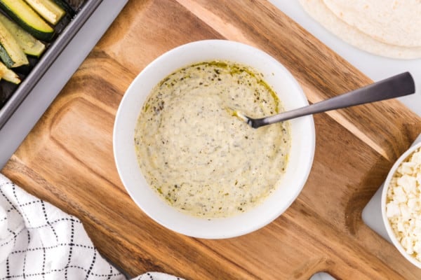 Pesto mayo in a white bowl with a spoon on a wooden cutting board.