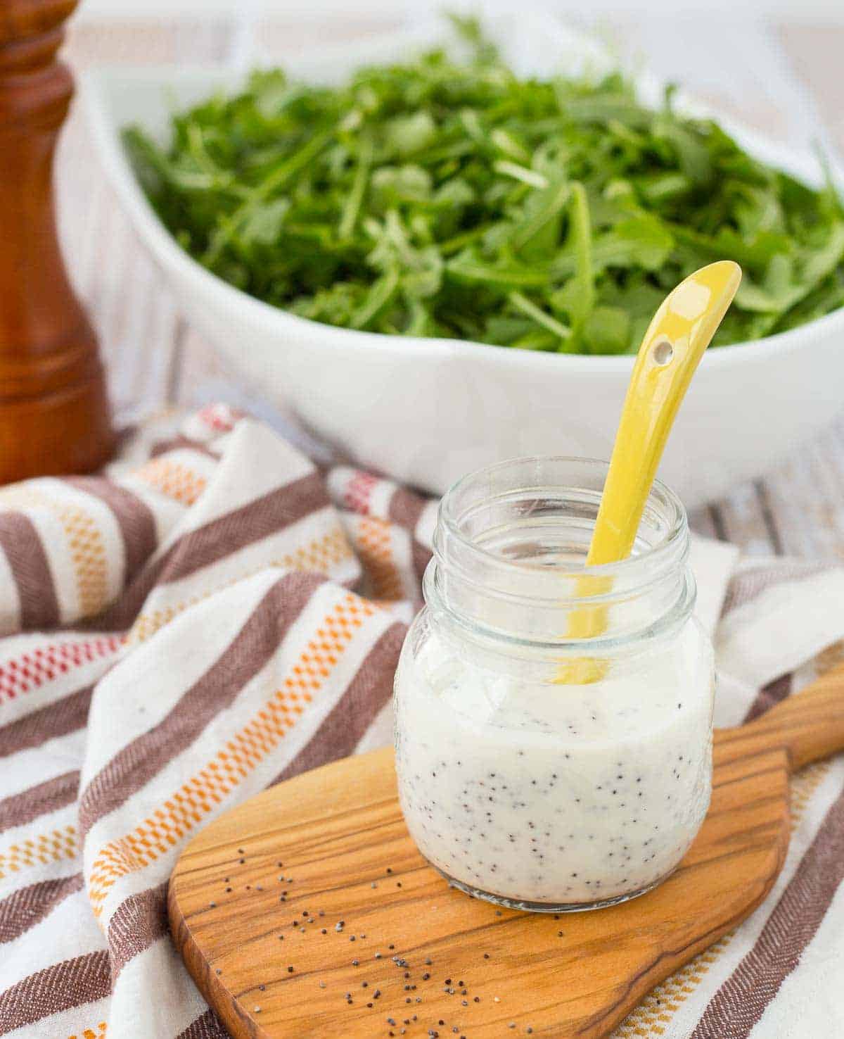 Front view of small glass jar with dressing, yellow spoon inserted, on decorative wooden board, with white bowl of arugula in. background.