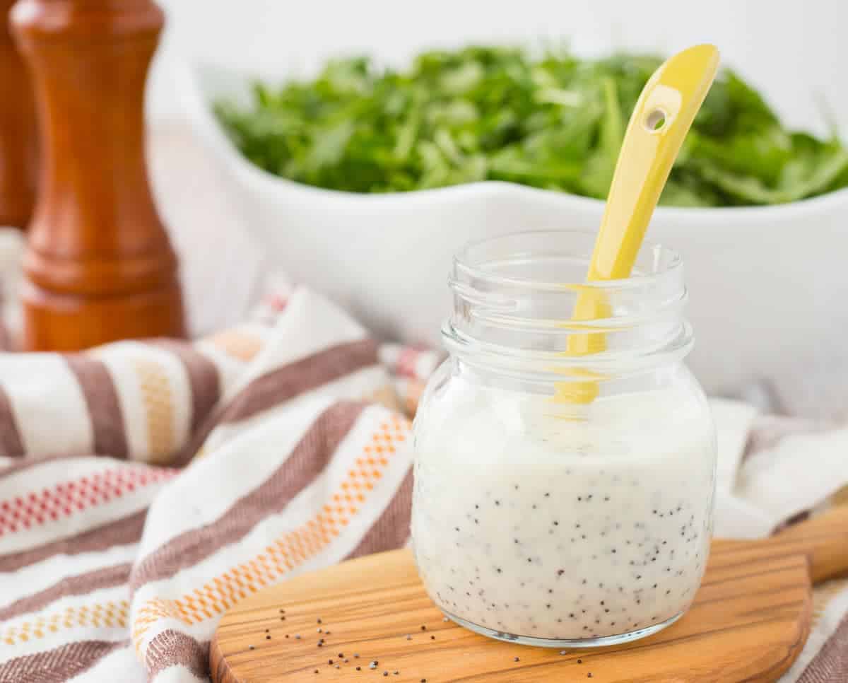 Front view of small glass jar with dressing and yellow spoon, with bowl of greens in background.