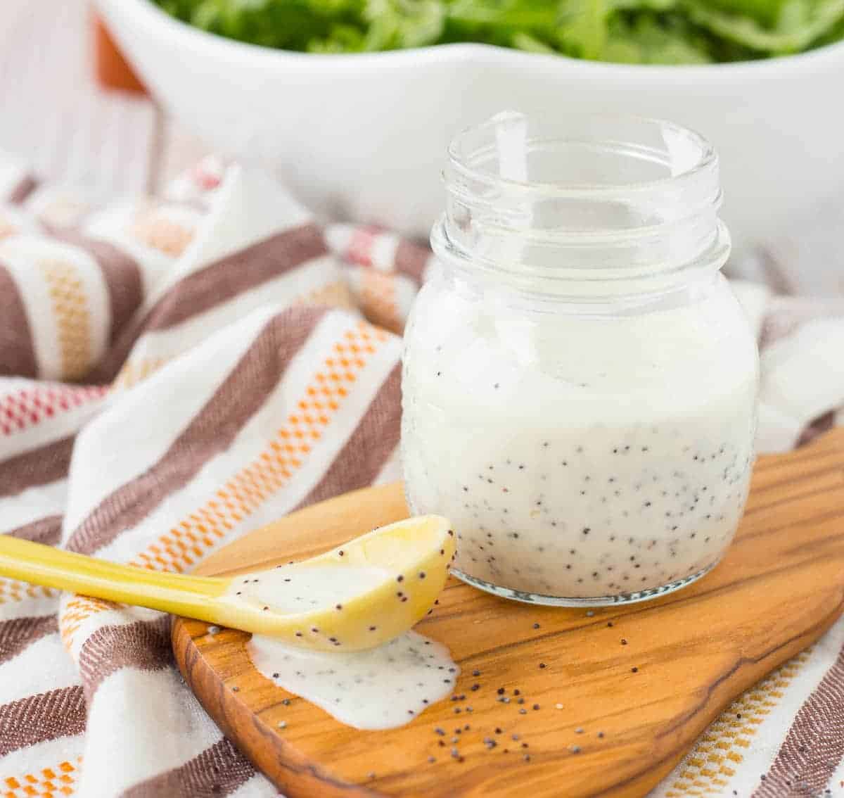 Small glass jar with dressing, spoonful along side with dressing running onto wooden cutting board.